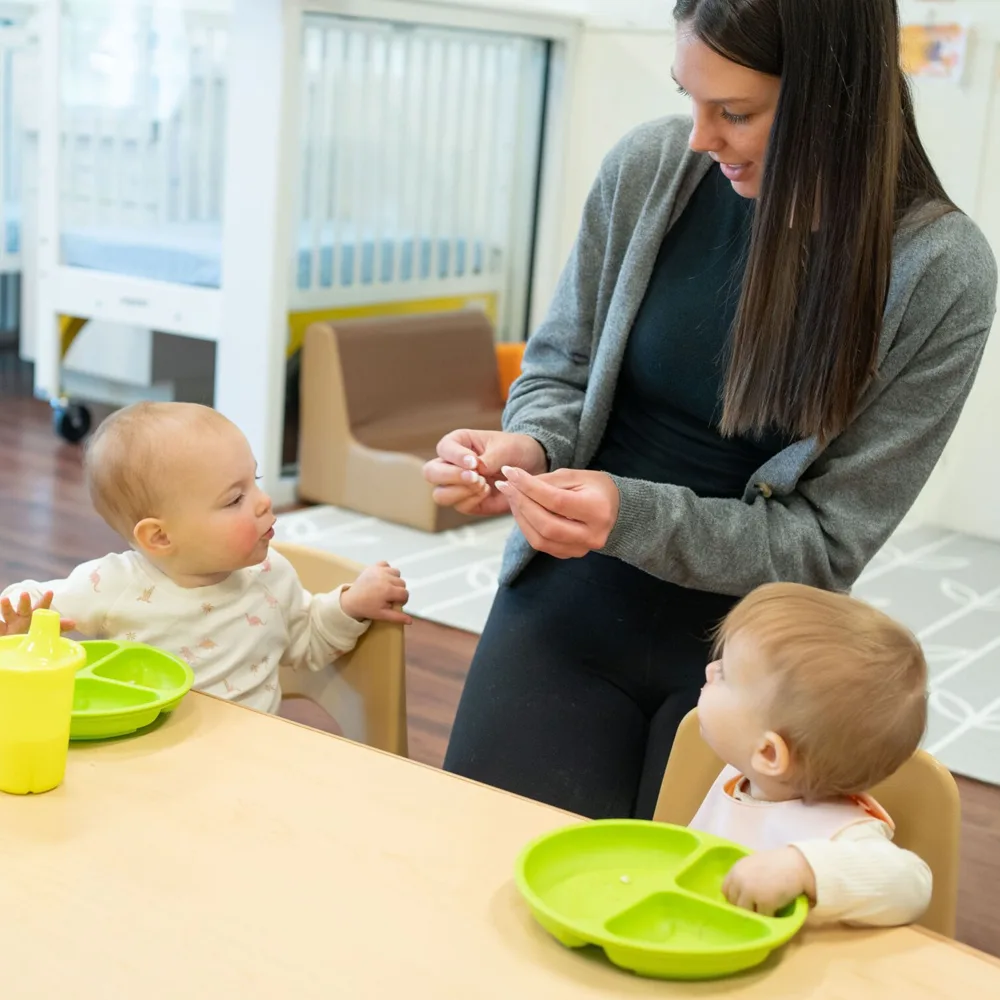 A teacher feeding two small toddlers at a table at a Peace of Mind daycare